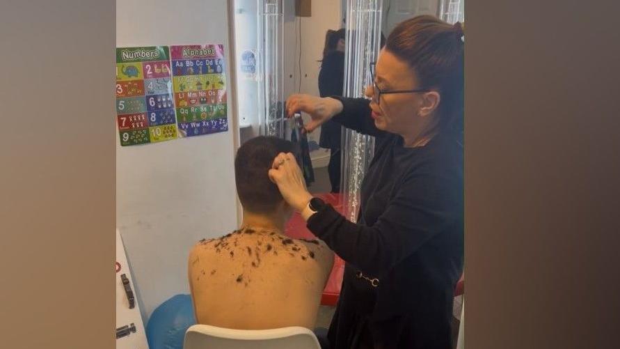 A photo taken from behind of a young boy sitting in a white chair while a woman cuts his hair.