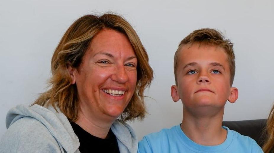 A woman wearing a grey hooded top over a black T-shirt. Next to her is a young boy wearing a blue T-shirt. He is looking upwards while the woman is smiling and looking into the camera.