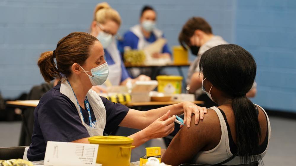 A nurse administers a vaccine does to a girl who is seated and faces away from the camera. The rest of the image is blurred but others can be seen also receiving their dose (file photo from March 2026)