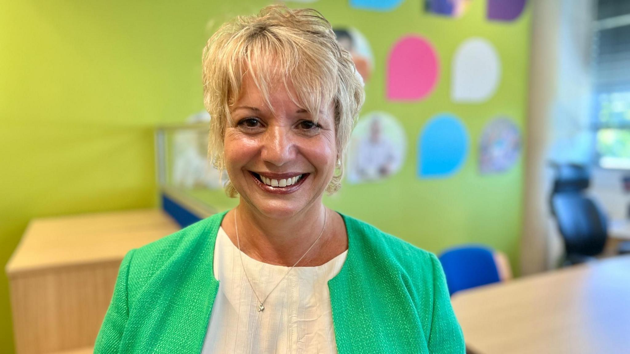 Sam Robinson looks at the camera wearing a bright green jacket. She has a white top on underneath it. She has short, blonde hair and is wearing earrings. She is smiling and is standing in the Big C office which is bright with a lime green wall and different colour teardrops showing photographs of people on the wall.