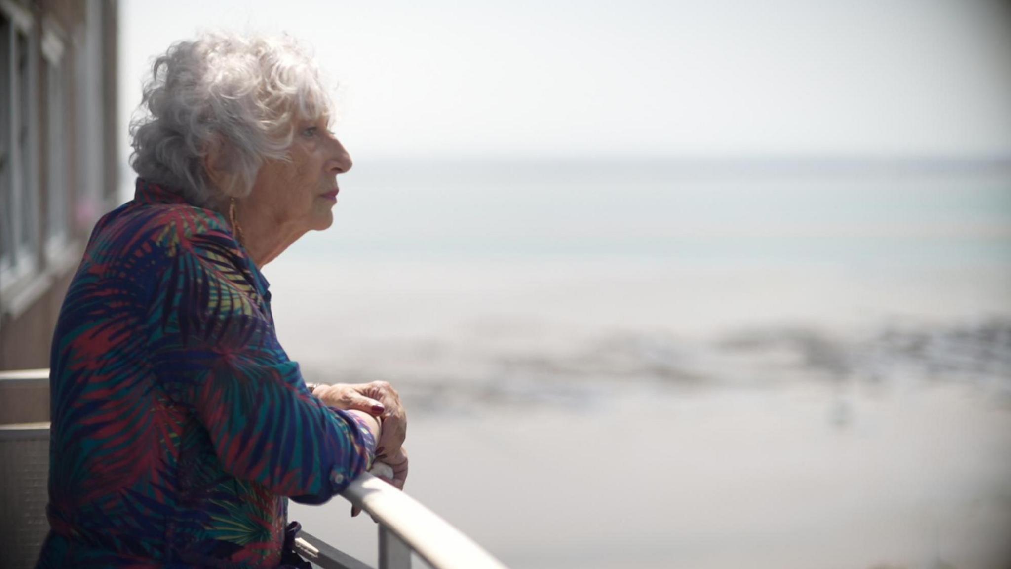 Carol Turansky standing on a balcony. She is wearing a leaf print dress and you can see her side profile. She has grey permed short hair and you can see the sea in the background. She is looking out to the horizon.