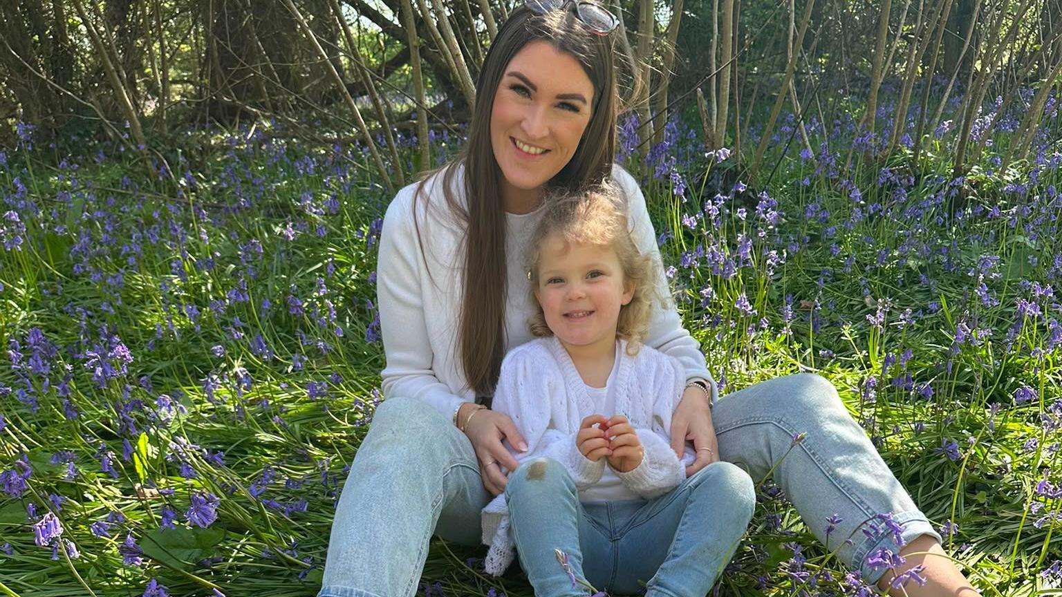 Alexandra is sitting outside on the ground surrounded by bluebells with three- year-old Ivy in front of her. They are both wearing white tops and pale blue jeans and are looking into the camera and smiling.