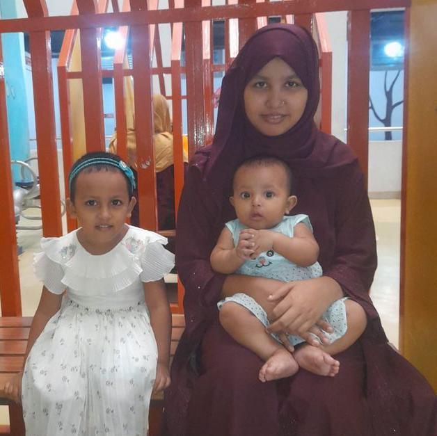 A woman wearing a maroon outfit sits on a wooden bench indoors, holding a baby in a light blue outfit, while a young child in a white dress sits beside them. Behind the bench are vertical orange bars and colourful wall decorations.