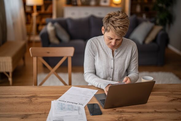 A woman sits at a wooden table in her living room. She looks at tax papers while typing on her laptop. A mug sits beside her and