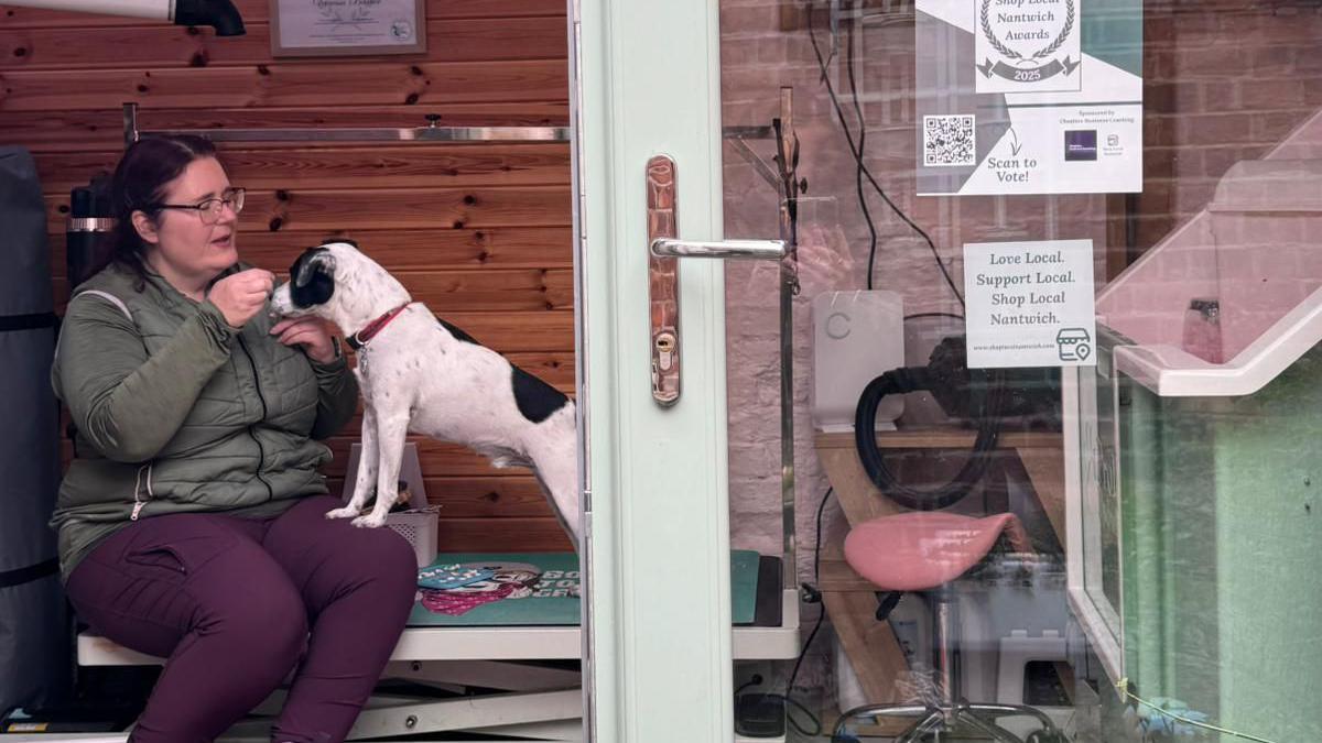 The shop window of a pet grooming business, showing a woman sat inside with a dog standing on her lap with his front paws