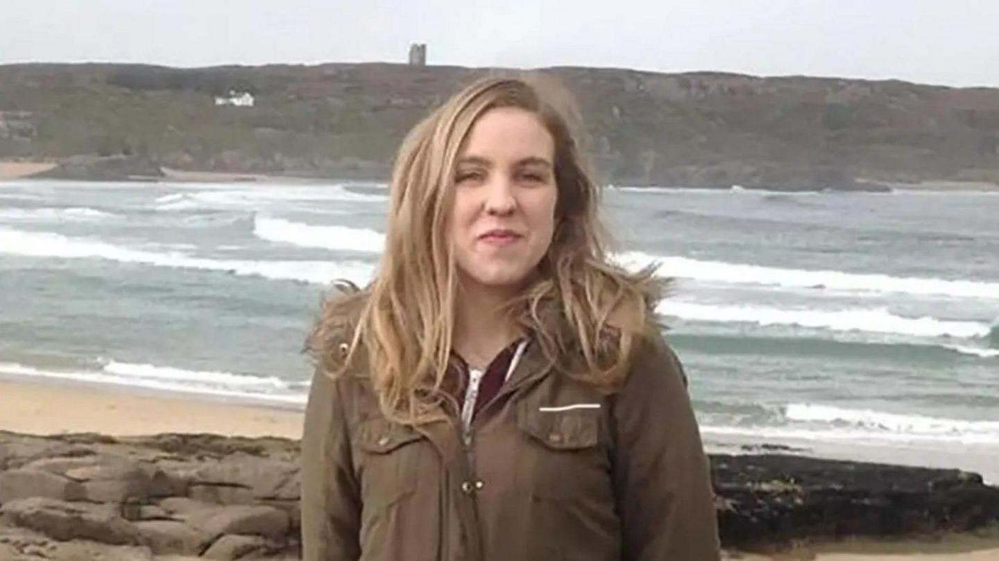 Natalie McNally standing on a beach with the sea visible behind her. She has long blonde hair and is wearing a dark green coat. She is smiling. 