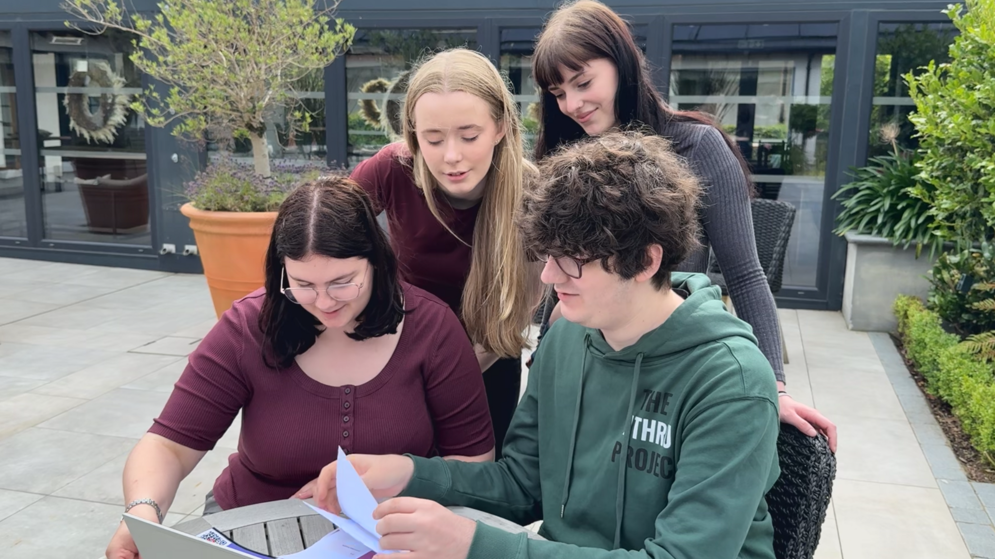 Four young people, two standing and two sitting, are looking at a laptop and some paper.