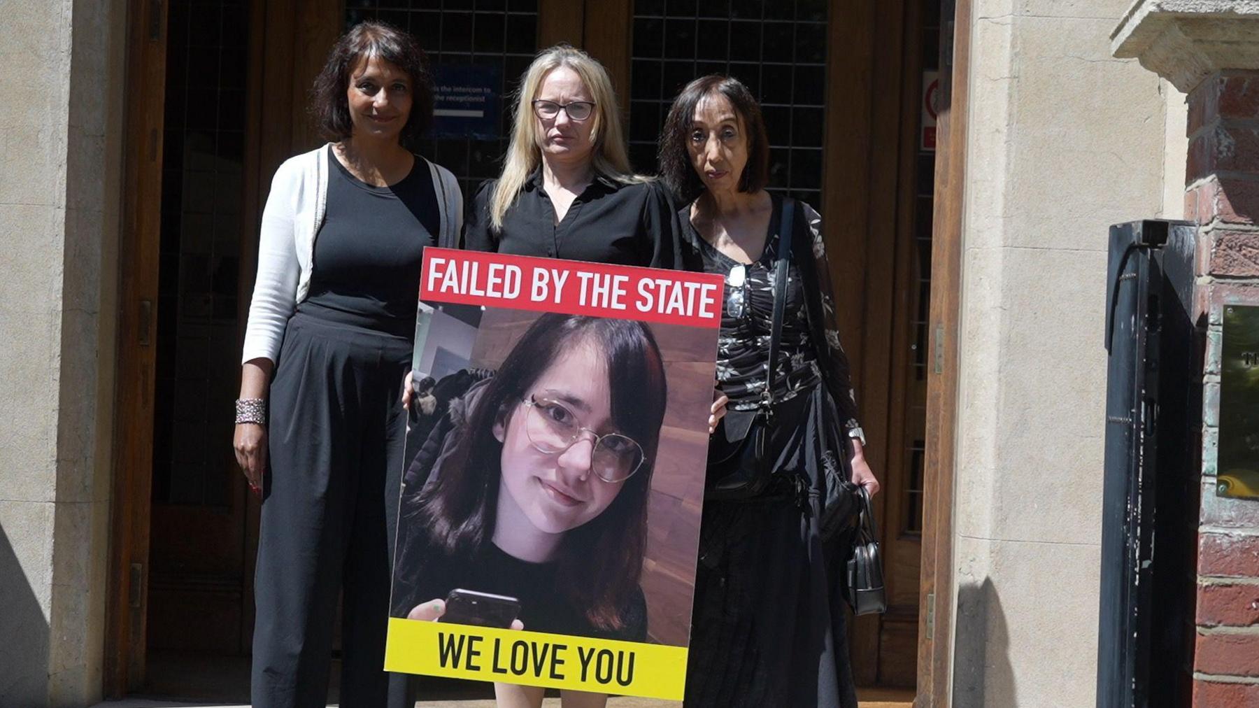 Victoria Sebastian stands outside the front of Arundel House in London holding a big poster of her daughter, with the words "Failed by the state" and "We love you". She has two solicitors beside her. They are all wearing black.