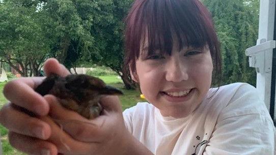 Elise Sebastian holding a small bird in her hand. She is smiling and is in a wooded area. She has a fringe and brown hair with a red tinge. She is wearing a white T-shirt.