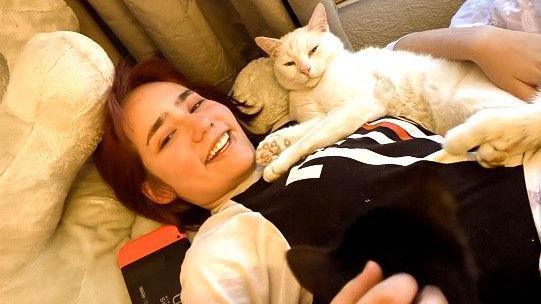 Elise Sebastian lies on a sofa with her cat on her. It is a white short haired cat, Elise is similing and wearing a long sleeved black and white top
