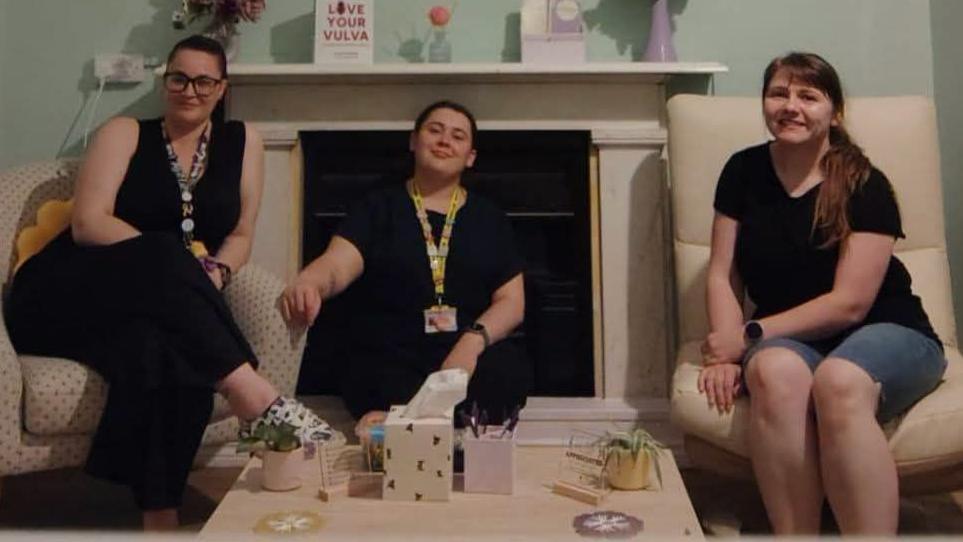 Three women wearing black sit in a living room.