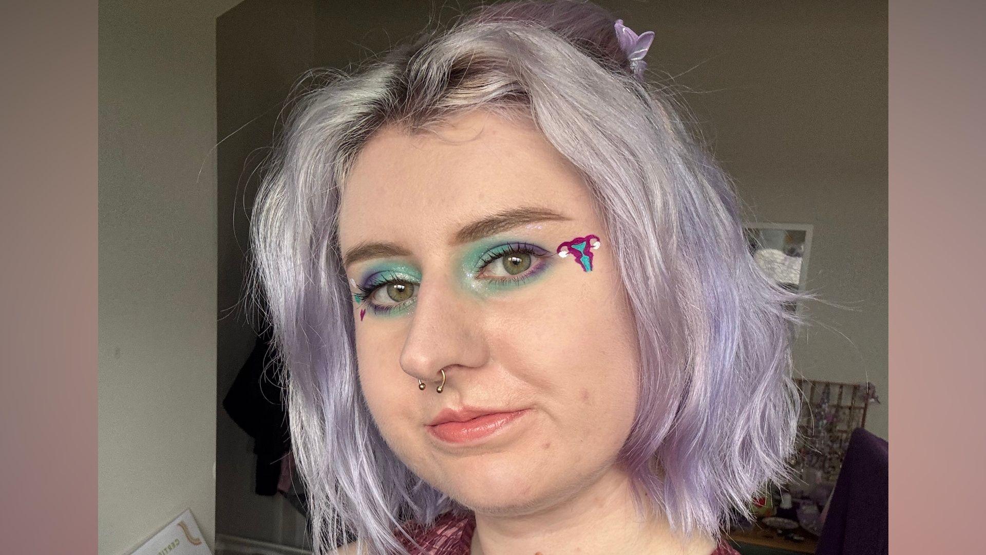 A headshot of a young woman with short light purple hair. She has blue and purple eyeshadow with a uterus-shaped sticker in the corner of her eye, and and a silver septum nose piercing.