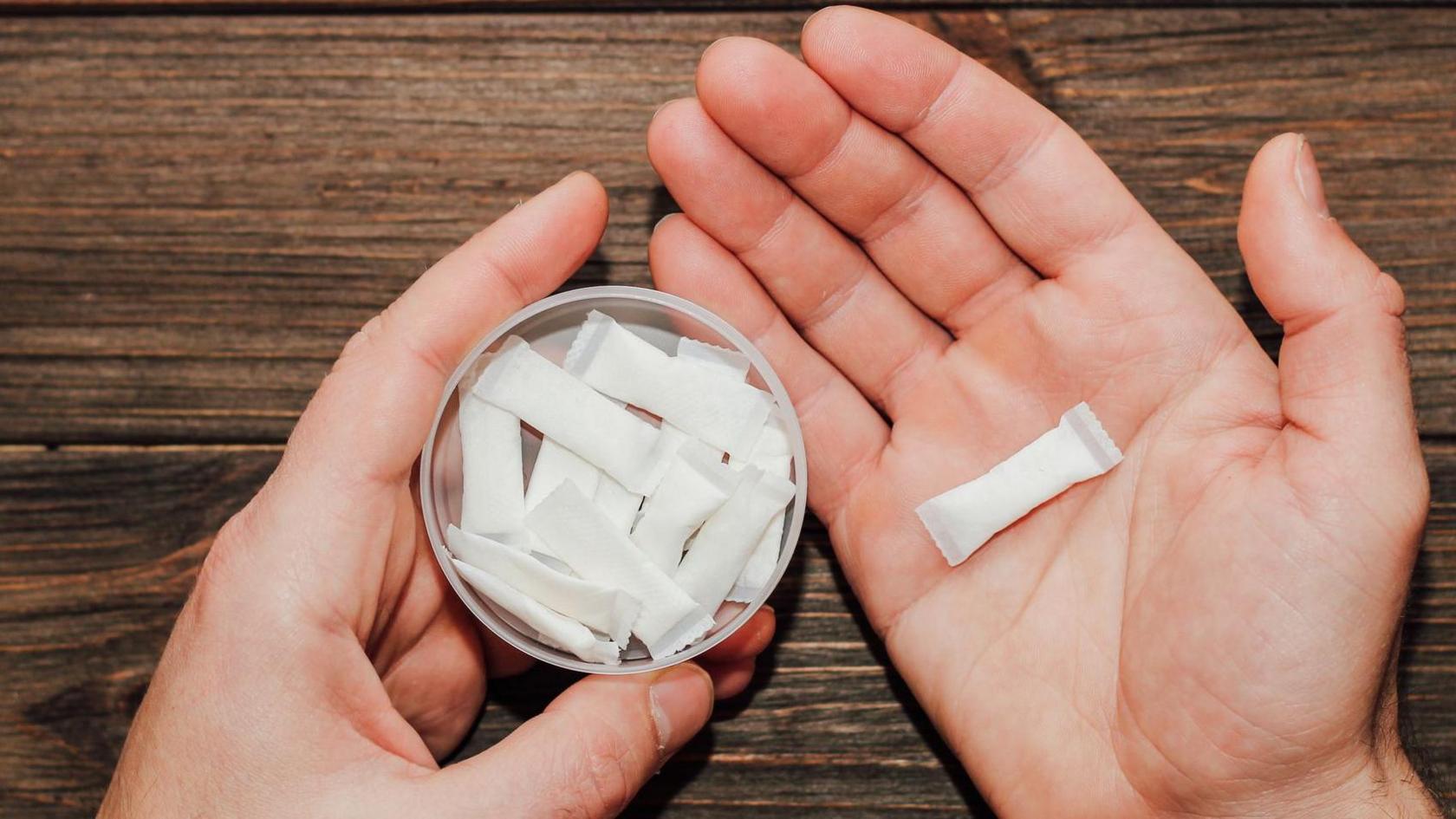 A pair of hands holding a can of pouches in one hand and a single pouch in the other. Pouches are white sachets.