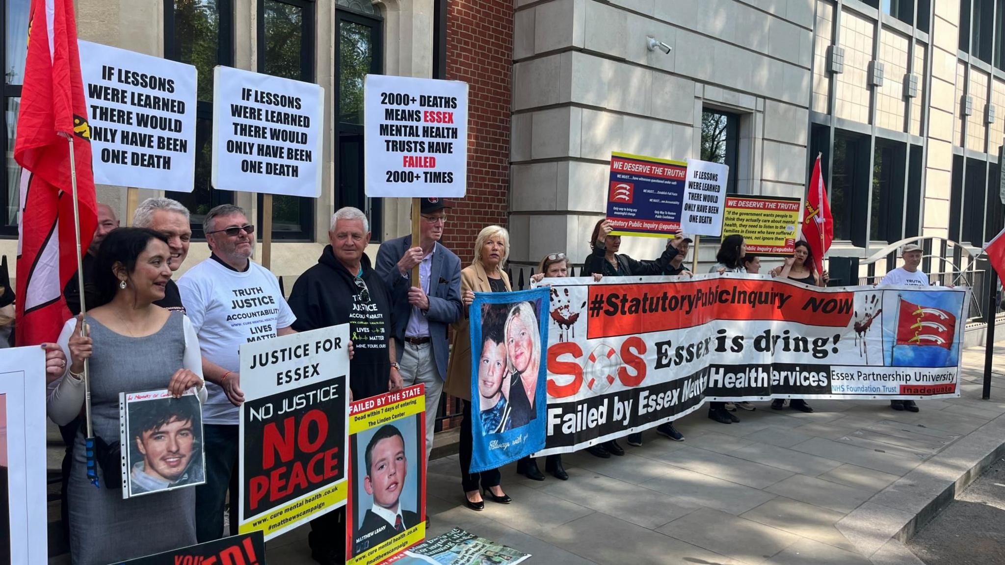 Protesters stand outside Arundel House in Central London. They are holding a big horizontal banner that says "SOS Essex is dying!" Some are also holding posters of their relatives that have died. They are standing outside the front of the mock-Tudor building. There is a row of about 30 people.