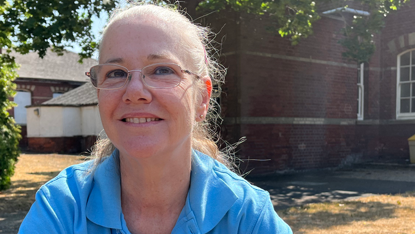 A woman with long blonde hair tied back is sitting on a wooden bench. She is wearing a blue polo shirt. She wears glasses.