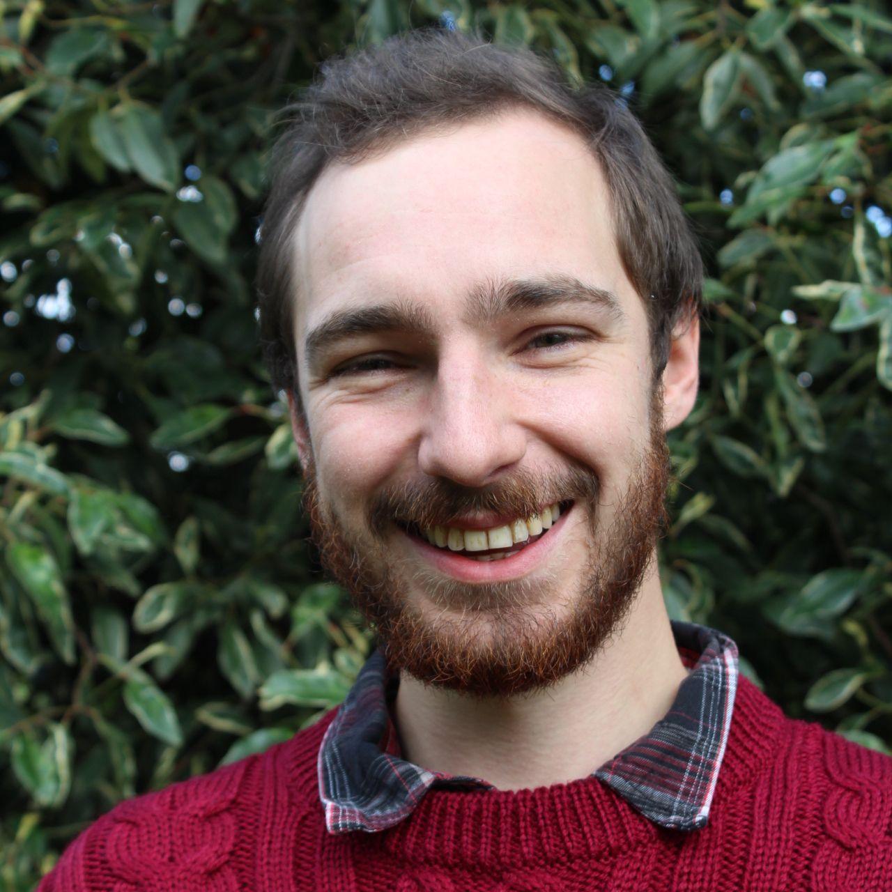 Headshot of young man with short brown hair and beard. He wears a checked shirt under a red jumper and is grinning at the camera.