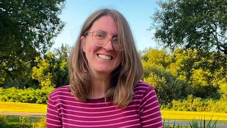 Head and shoulders shot of young woman with glasses and shoulder-length light brown hair. She wears a pink striped top and is surrounded by countryside.