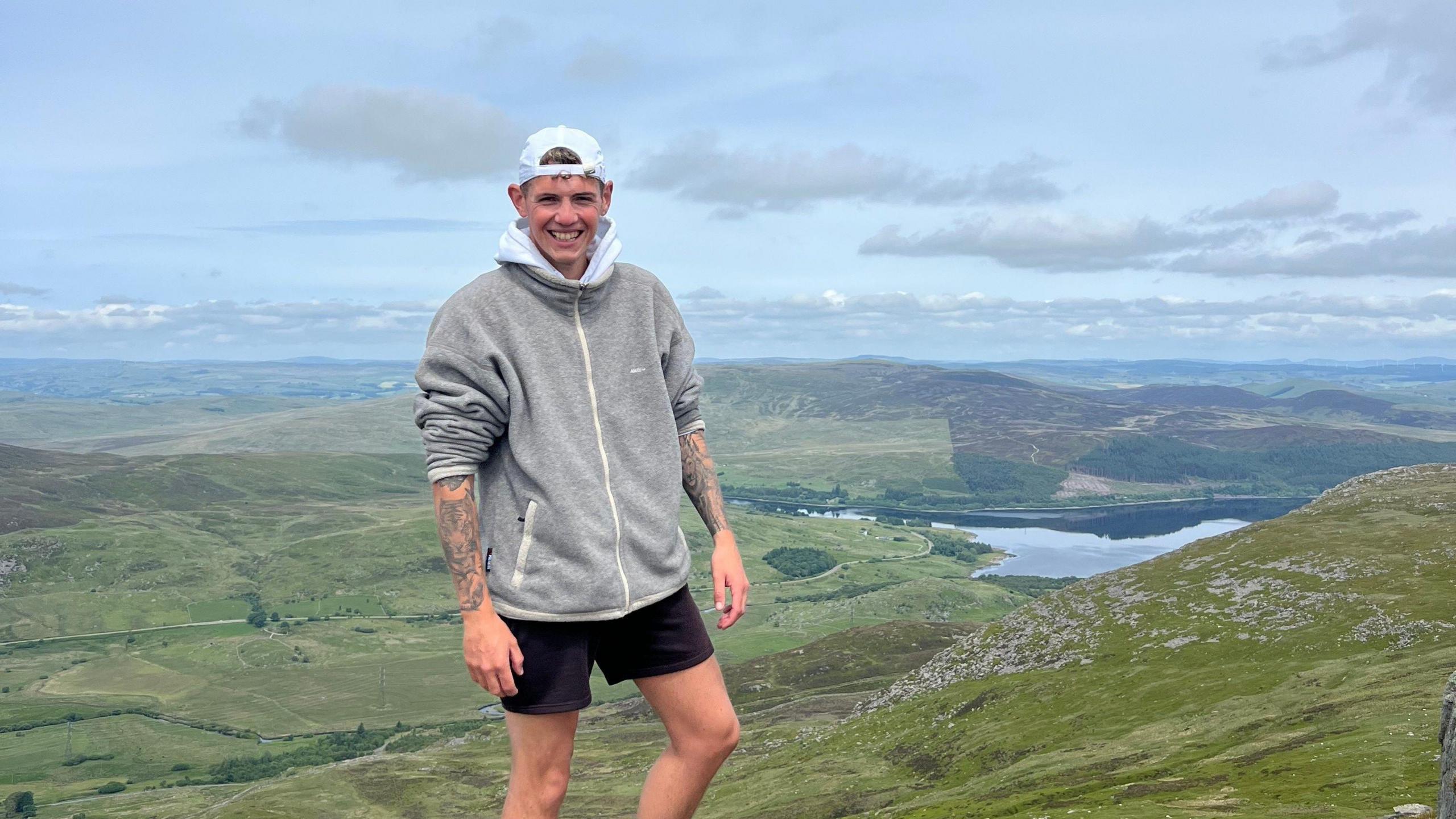 Ed Shirt wearing a hoodie and shorts standing on a mountain with a view behind him