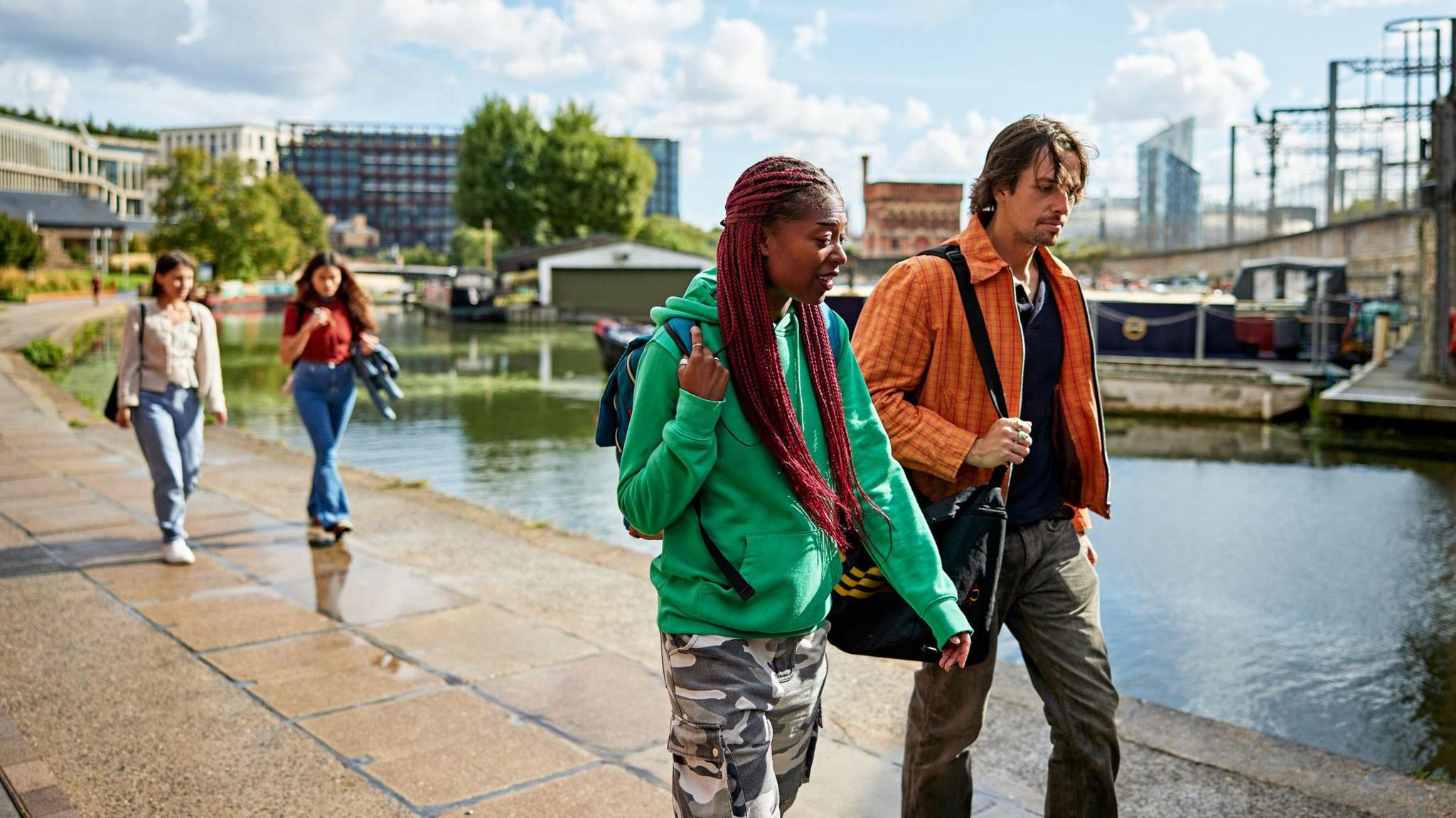 Four casually dressed friends in 20s and 30s, two walking in front and other two behind them, enjoying stroll along canal 