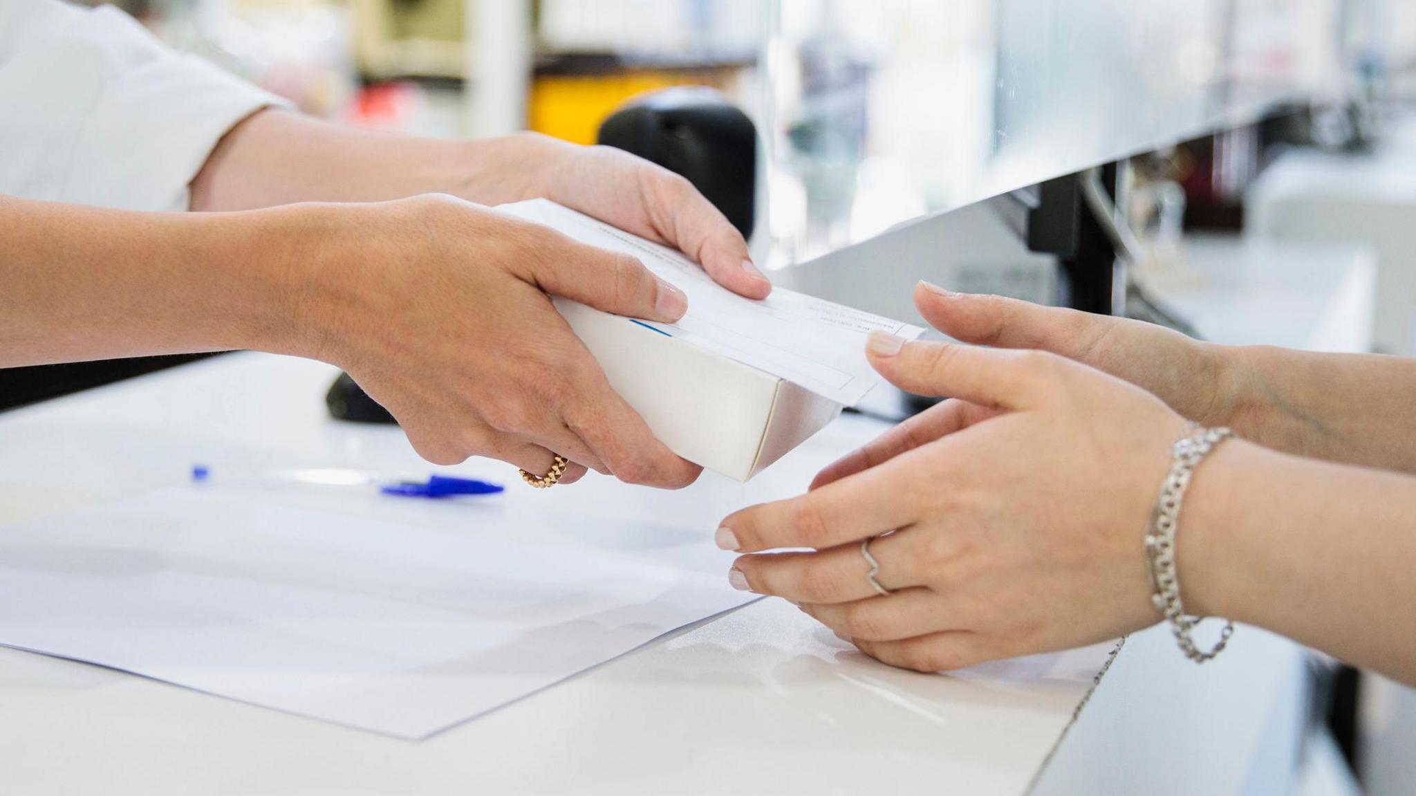 Two sets of hands are visible in the picture, with one person handing a white box to another person. The background make it obvious they are in a chemist. 