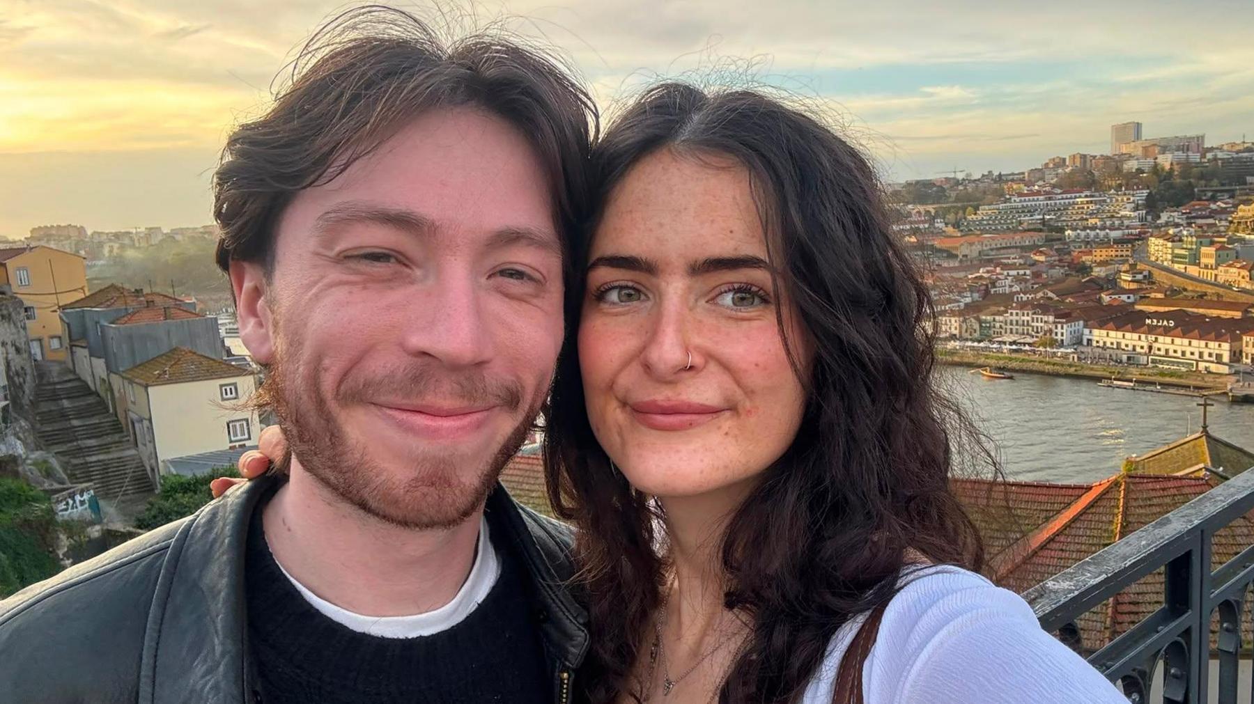 A smiling young couple with dark hair take a selfie at a scenic viewpoint, with buildings and water visible in the background.