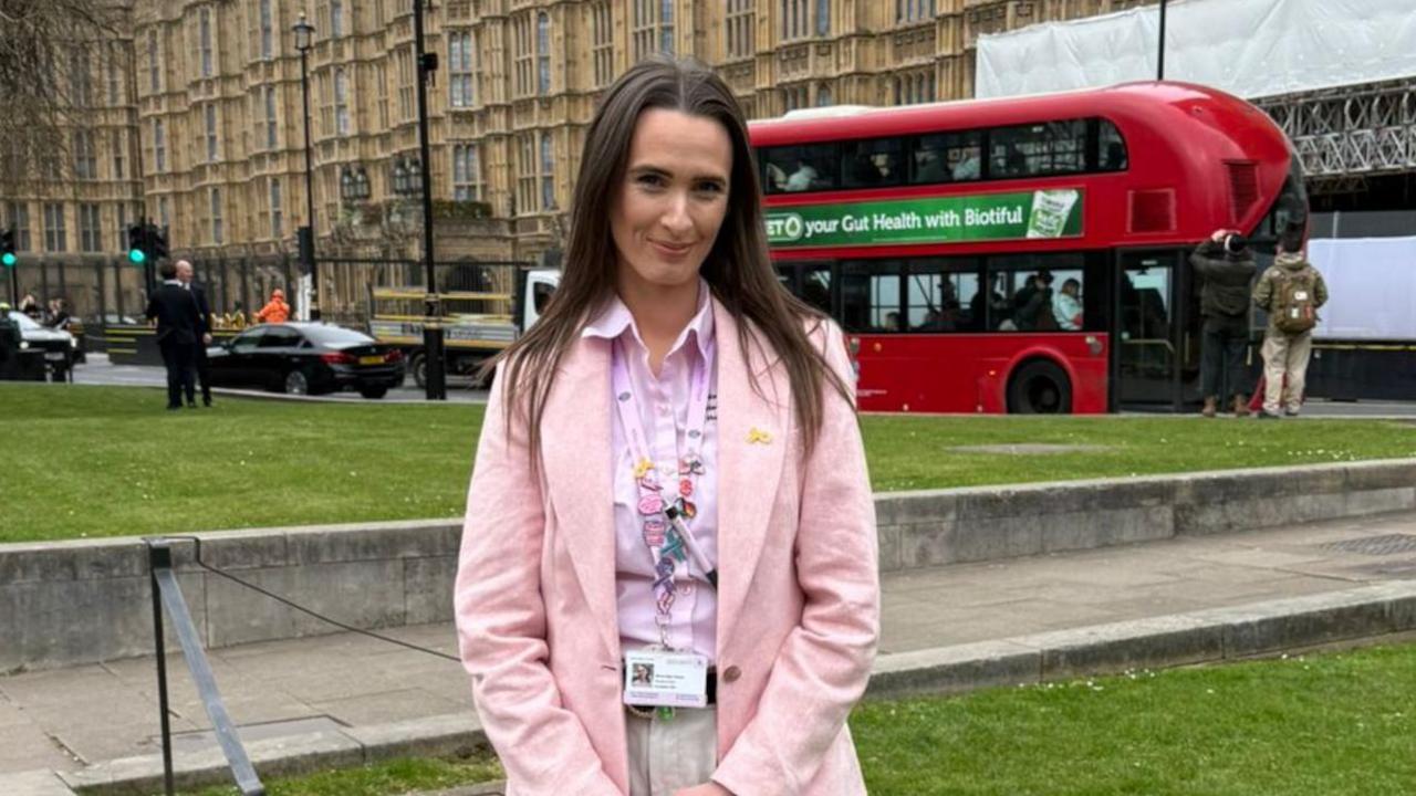 A woman stands outside of Westminster in London. She has long brown hair and wears a pink blazer with a pink shirt and cream trousers. Vehicles pass along a road behind her.