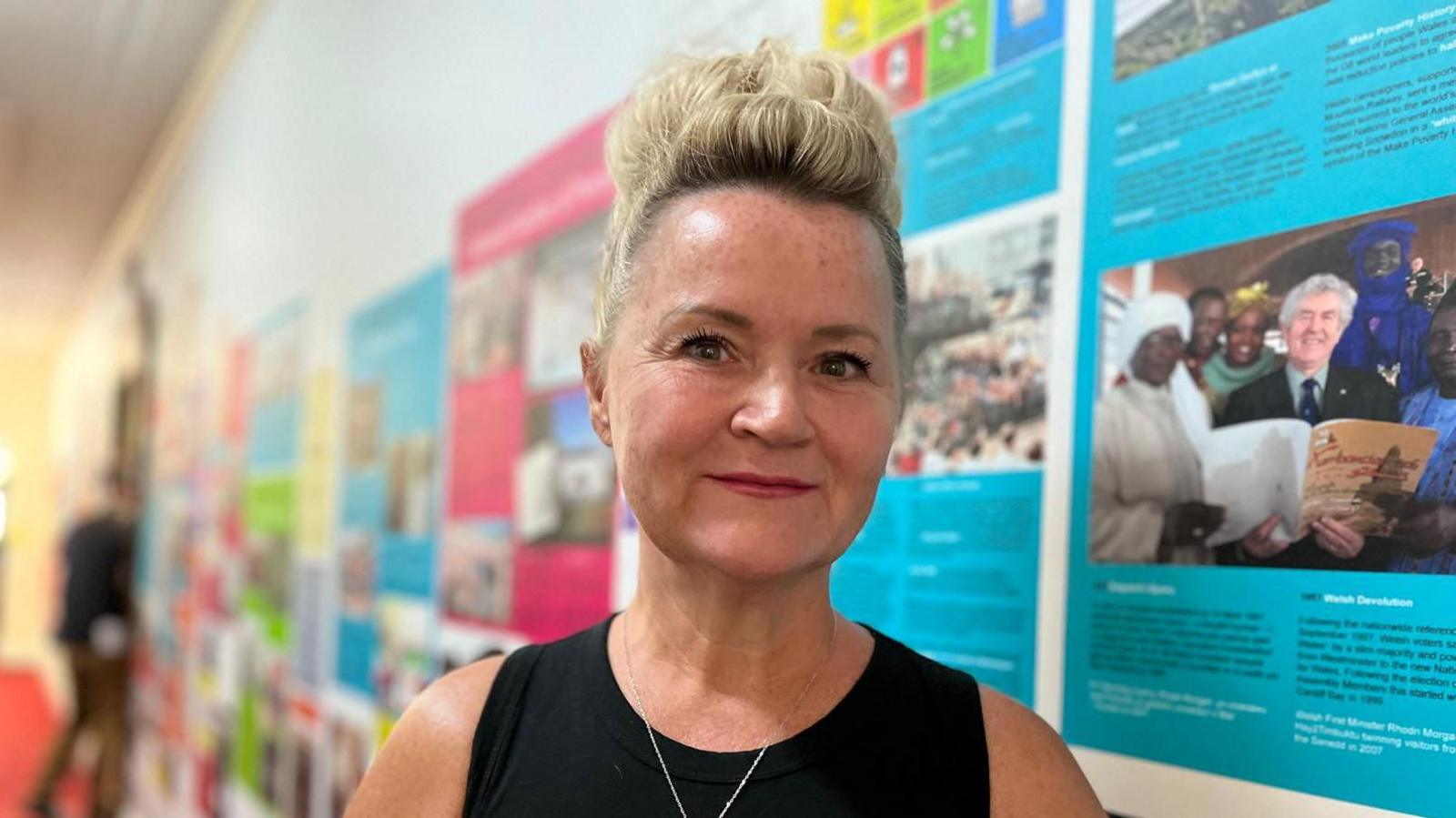Donna is wearing a black sleeveless top and is smiling at the camera. She is stood in a corridor with images and articles in bright colours along the wall. She has short blonde hair which is shaved on the side and has a quiff on the top