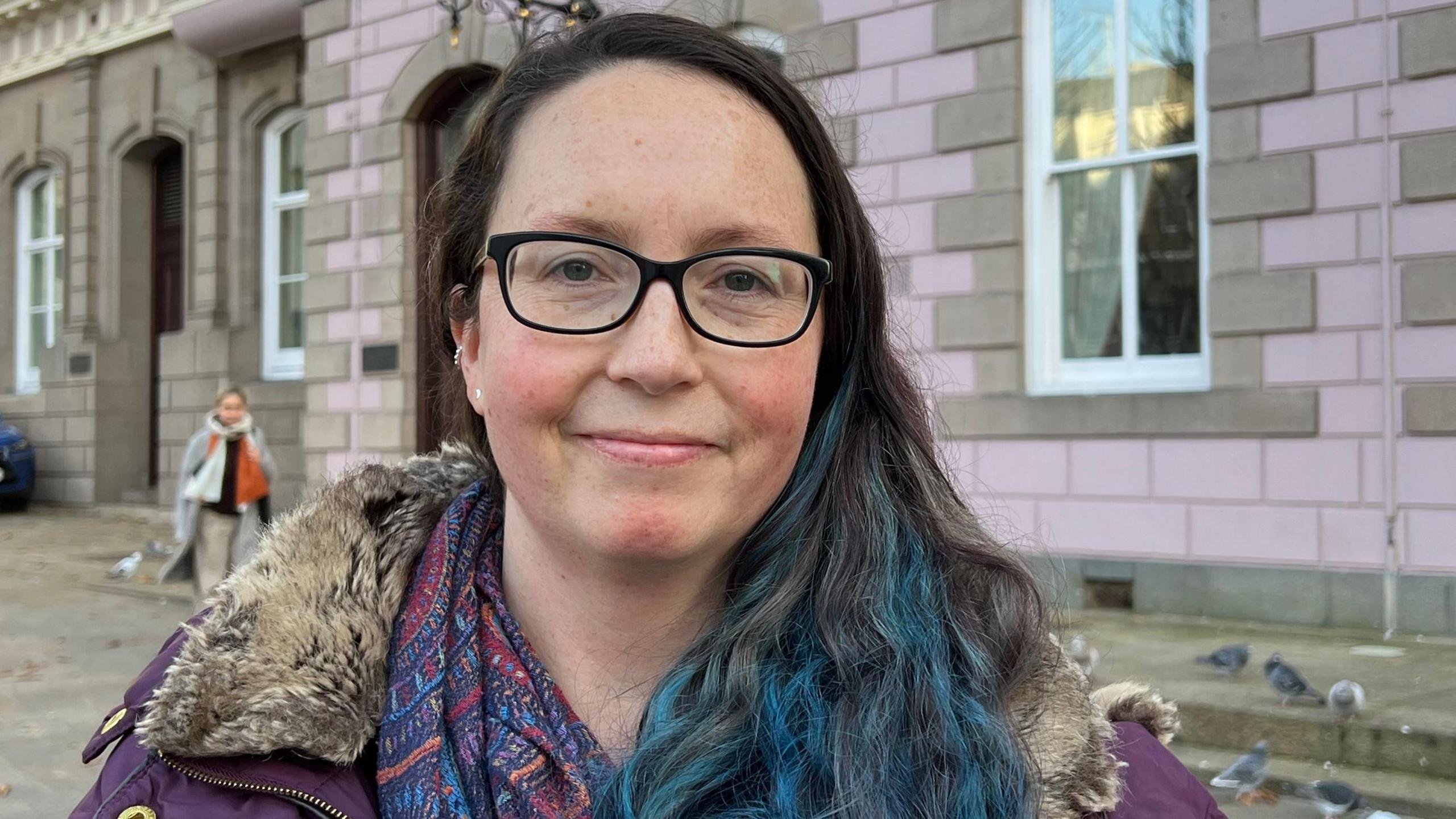 Deputy Louise Doublet in Jersey's Royal Square. She has long dark hair, with blue streaks at the ends. She wears glasses, a purple coat and a patterned scarf.