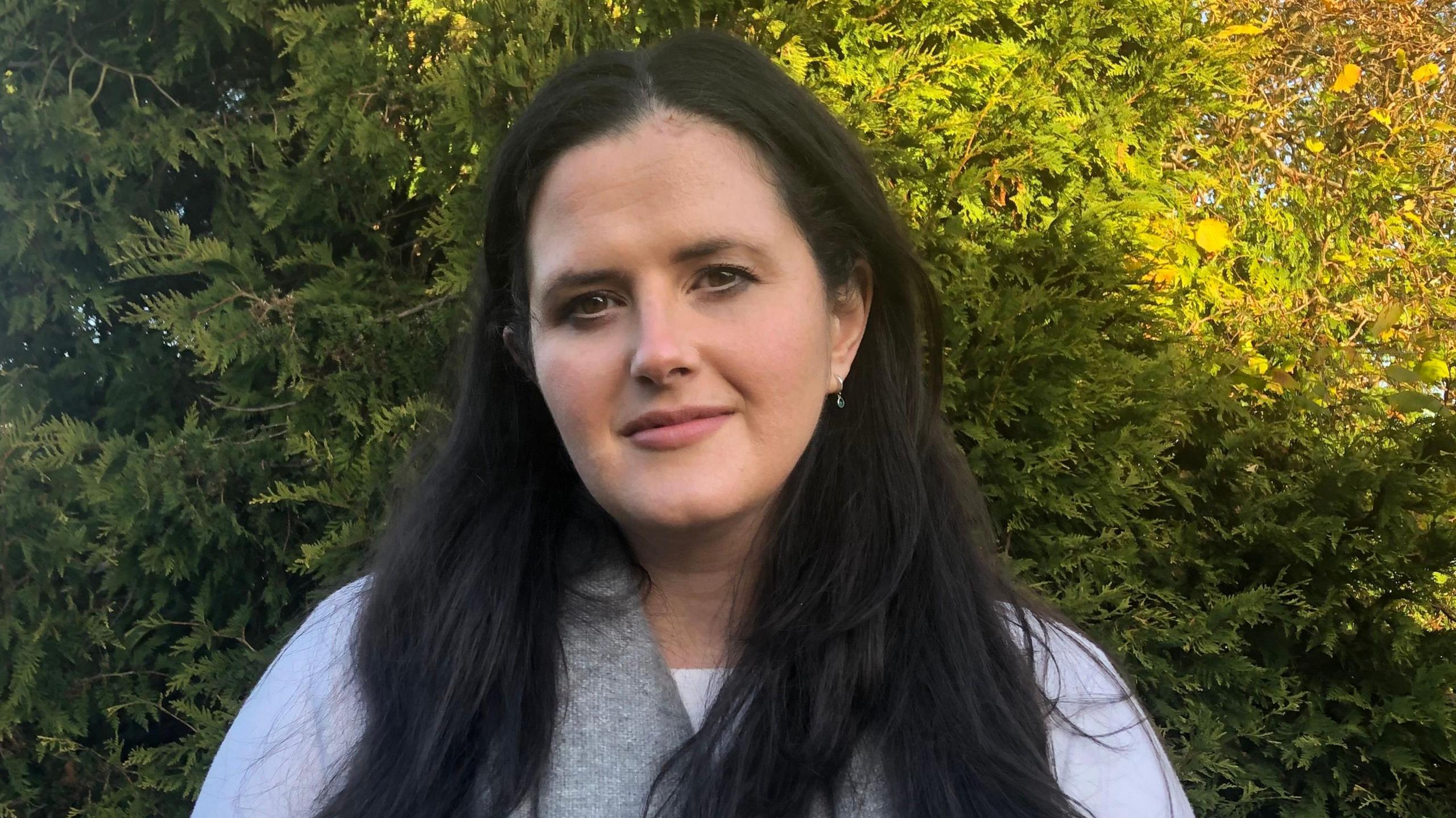 Claire Sugden, a woman with long, straight black hair, looks to the camera while standing outdoors on a sunny day. She is wearing a white long-sleeved top and a grey scarf. There are evergreen trees in the background.
