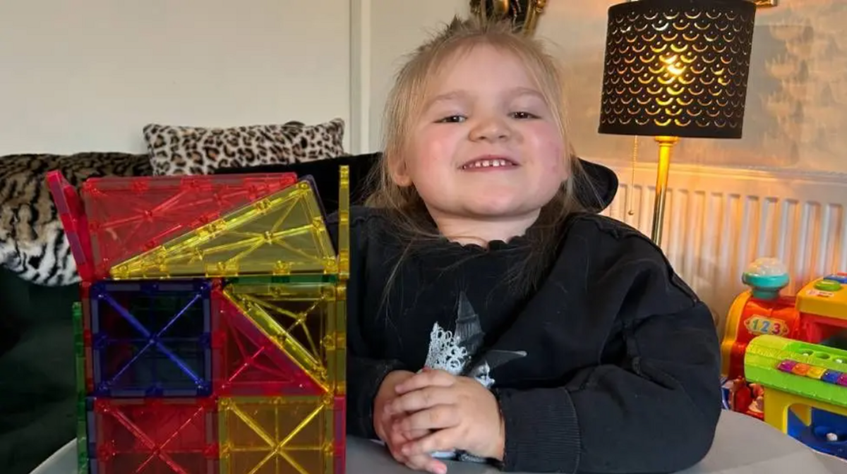 Dani sits at a table in a lounge, with toys in front of her. She smiles at the camera.