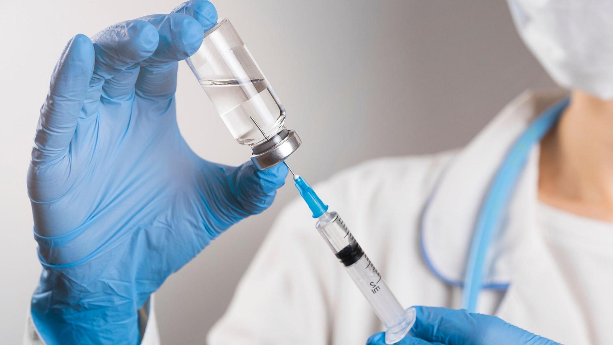A doctor in a white coat and blue protective gloves holds a medicine in a syringe. The hand of the medical worker knocks on the syringe and knocks out the air with his finger.