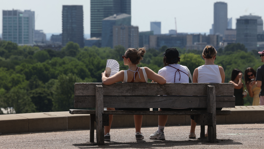 Three people sit on a bench in the sunshine as one woman fans herself from the heat