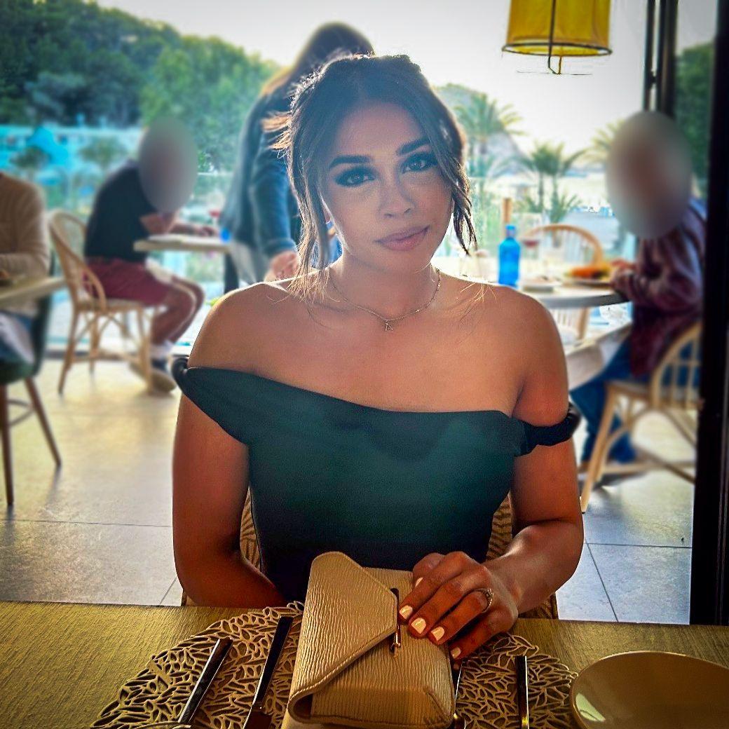 A photo of a young woman sitting at a table in a busy restaurant. Her brunette hair is tied back and she is wearing an off-the-shoulder black top, with her nude-toned handbag on the table.