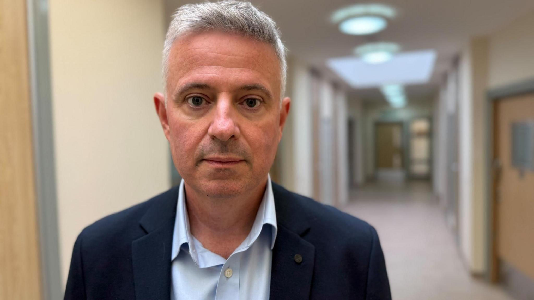 Paul Scott looks at the camera. He is standing in front of a corridor near the trust's headquarters. It is an inpatient unit. You can see a new beige corridor with light colured wood doors. Scott is wearing a navy suit jacket with a pale blue shirt