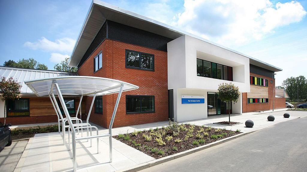 The front of the St Aubyn Centre, which is a red brick building with a cycle rack outside. There are rainbow-coloured windows to the side and it has a few plants in a bed at the front.
