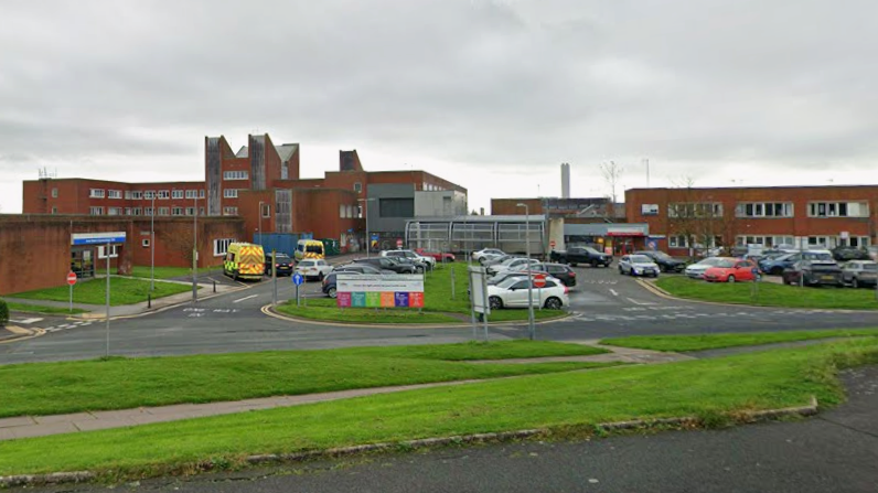 Furness General Hospital in Barrow. Buildings with orange bricks are interspersed with roads and grassy banks. Ambulances are parked to one side.