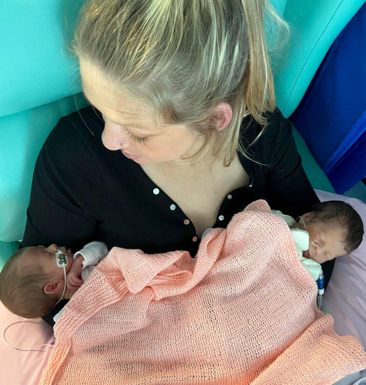 Brioney holds her newborn identical twin daughters in 2022 - they lie either side of her as she sits up in a chair, wearing a black top. The babies bodies are covered with an orange blanket.