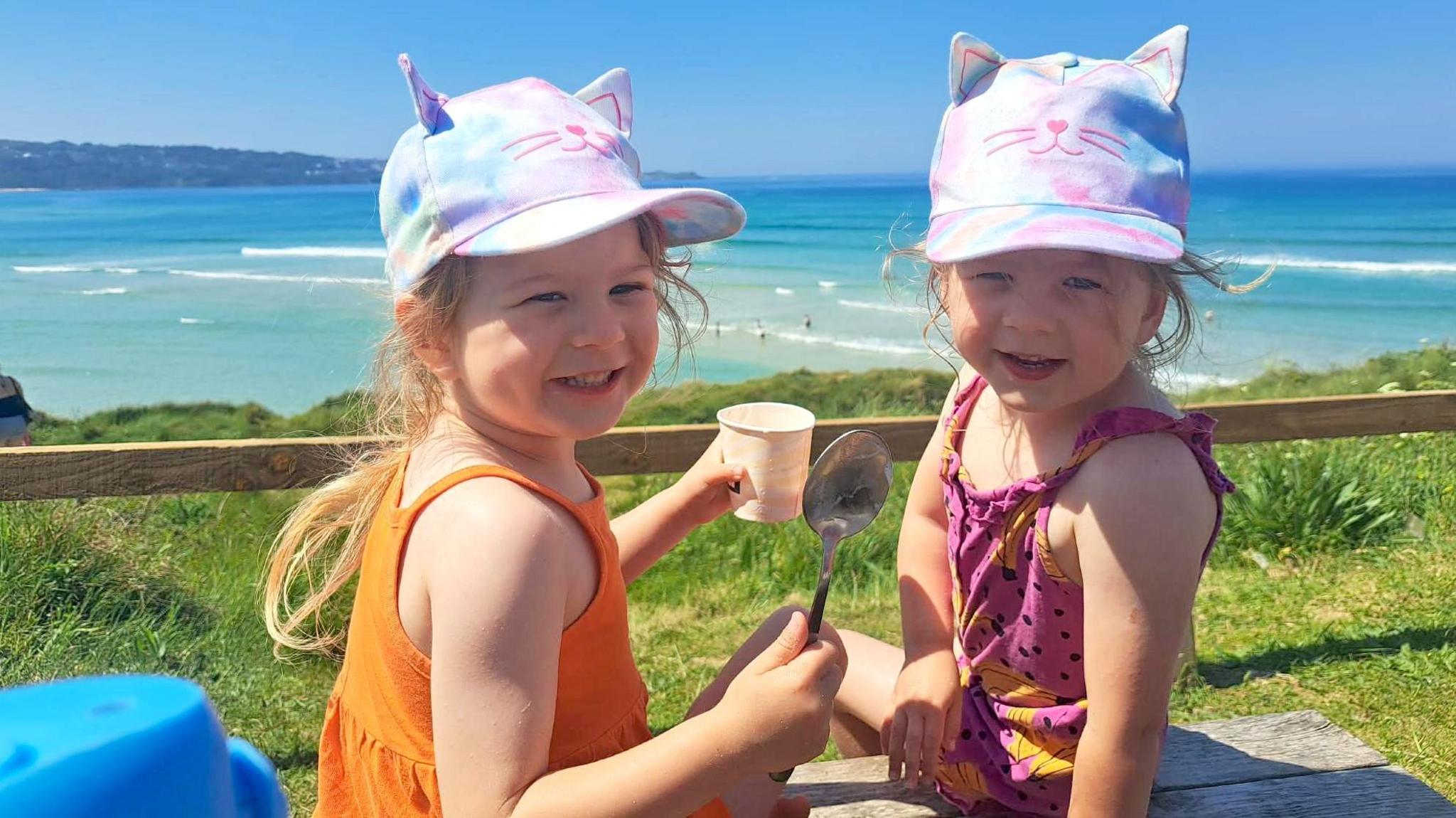 Identical twins Nancy and Margo eat ice cream at the beach, wearing colourful caps and strappy tops, with azure-blue sea and gentle waves visible in the background.