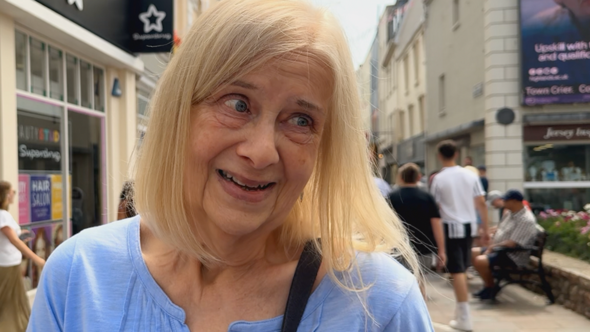 Interviewee Carole - a woman with shoulder-length light-coloured hair, wearing a light blue top and a cross-body bag. She is standing on a pedestrian Jersey's high street. Storefronts and signs line the street behind her, while several people walk or sit in the background. 