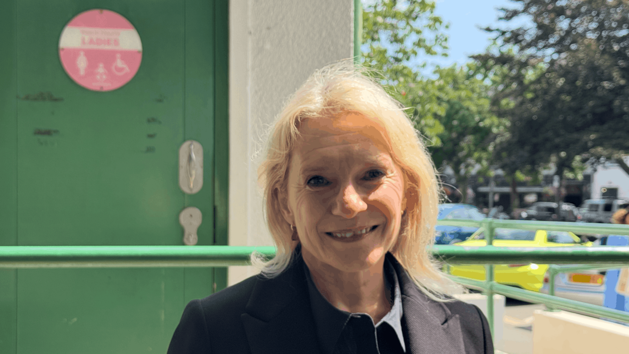 Senator Elaine Millar wearing a dark jacket, standing outside a public toilet entrance, with a green accessible restroom door visible in the background.