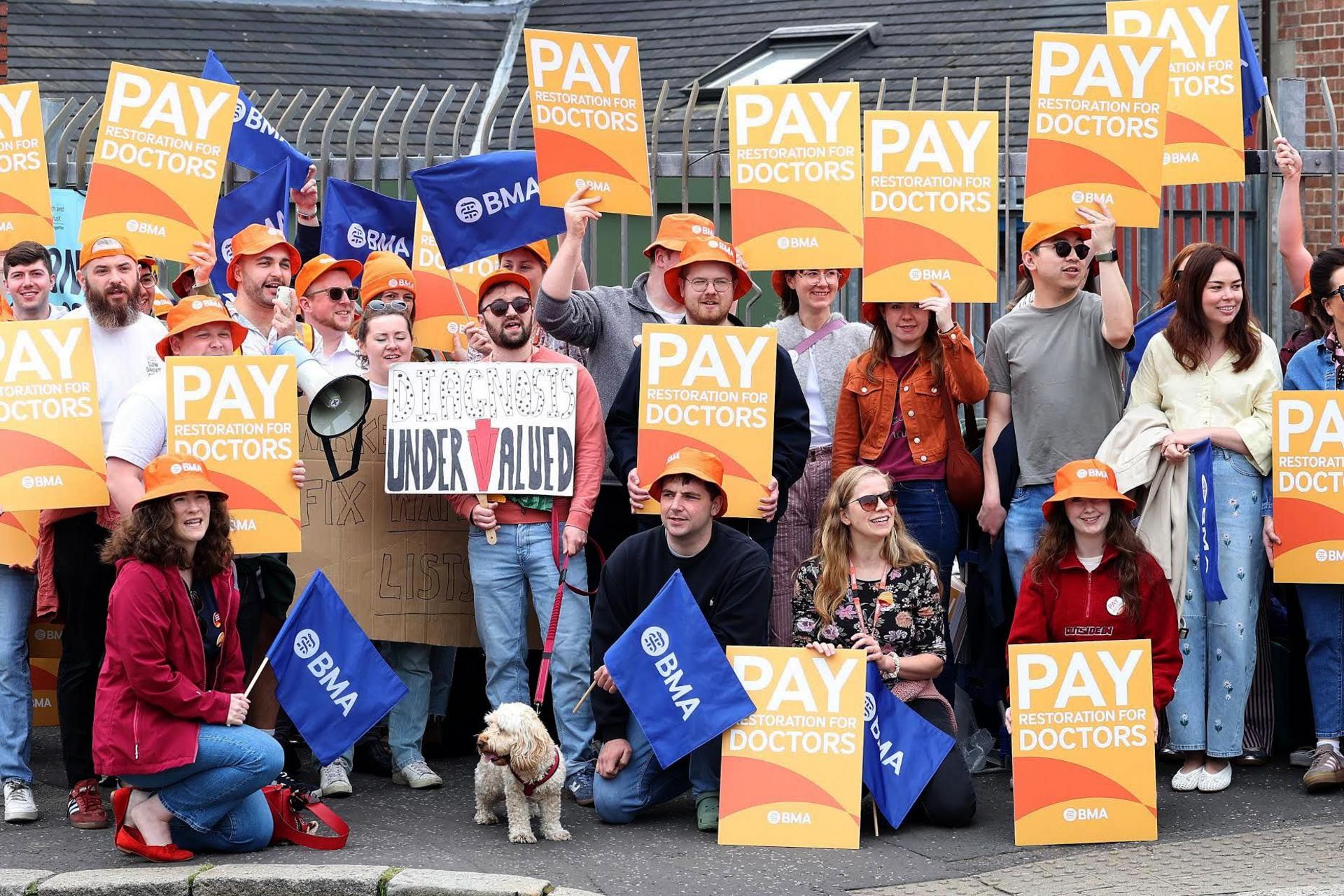 A large crowd of people on strike,, holding orange placards that read "pay restoration for doctors"