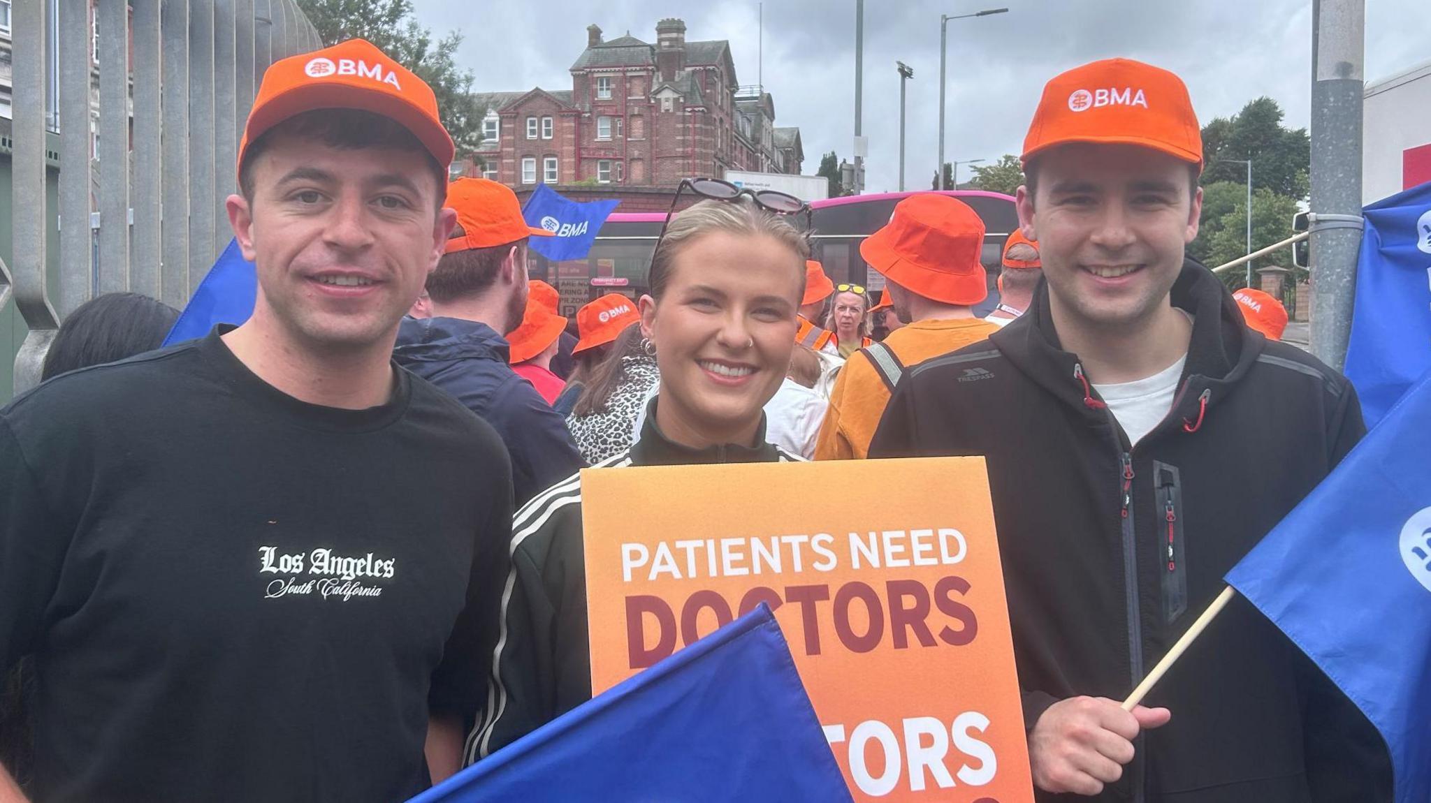 Orla Murray standing with two other protesters holding blue BMA flags and wearing orange hats dressed in black. Orla has blonde hair, is wearing glasses on her head as well as an adidas top