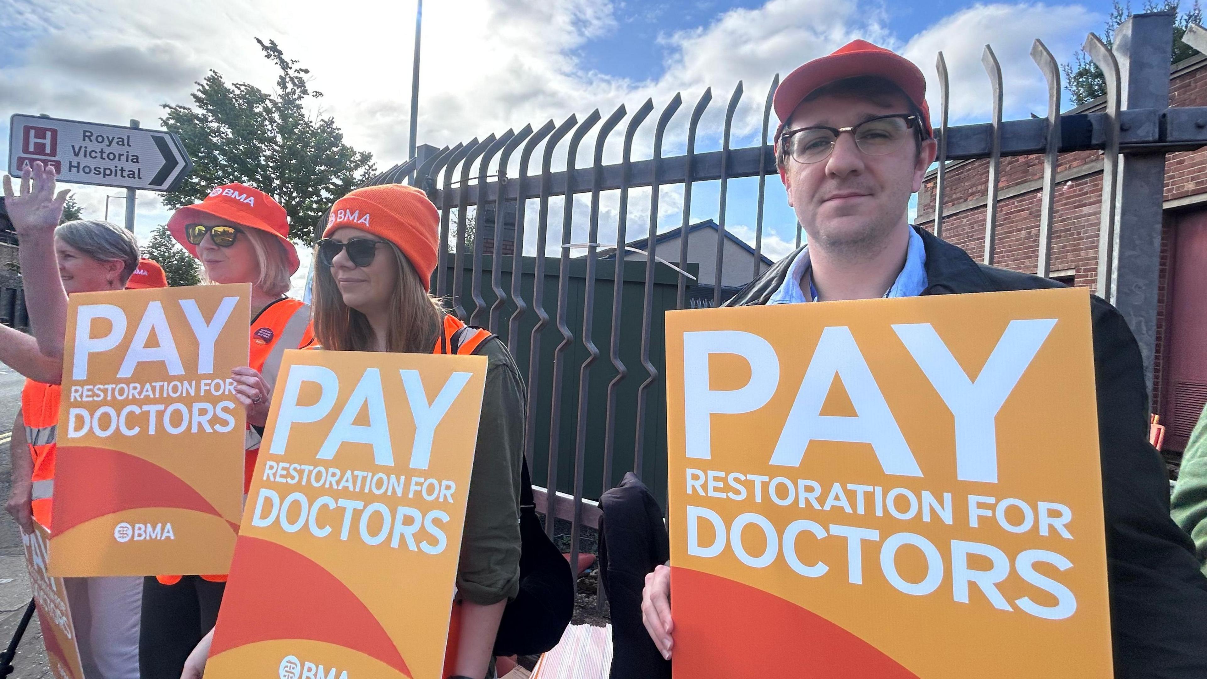 Doctors on strike standing outside a hospital with orange signs with 'Pay restoration for doctors' written on it.