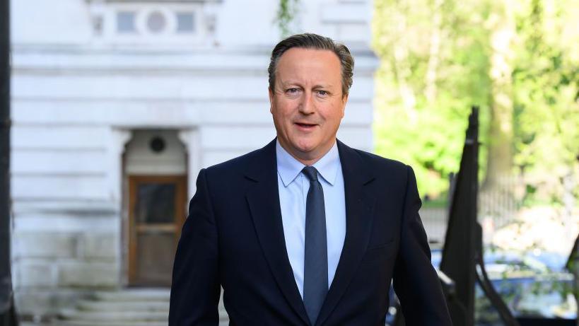 The former Prime Minister David Cameron is walking down a street looking at the camera, wearing a navy shirt, light blue shirt and mid blue tie. There is a white building with steps leading up to a brown wooden door in the background and some black railings to the right of the picture.