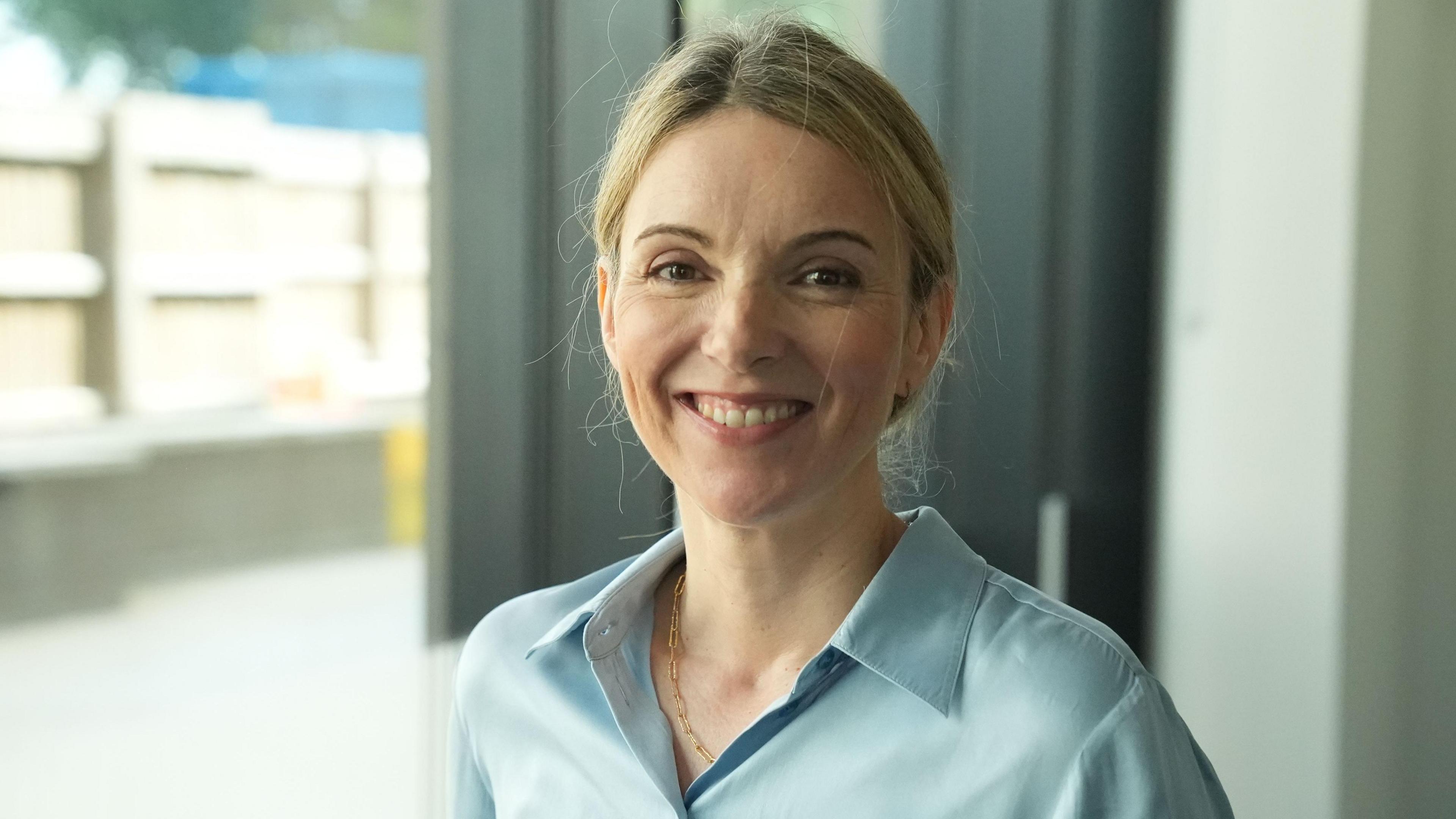 The image shows a person standing indoors near large windows in a bright, modern setting. The person is wearing a light blue button-up shirt and a thin necklace, with hair pulled back away from the face. The photograph is framed as a head-and-shoulders portrait, with the subject positioned prominently in the foreground. The background is softly blurred and includes window frames, a curtain, and an outdoor area visible through the glass. Natural light illuminates the scene, creating a clean and professional appearance with soft shadows and neutral colors.