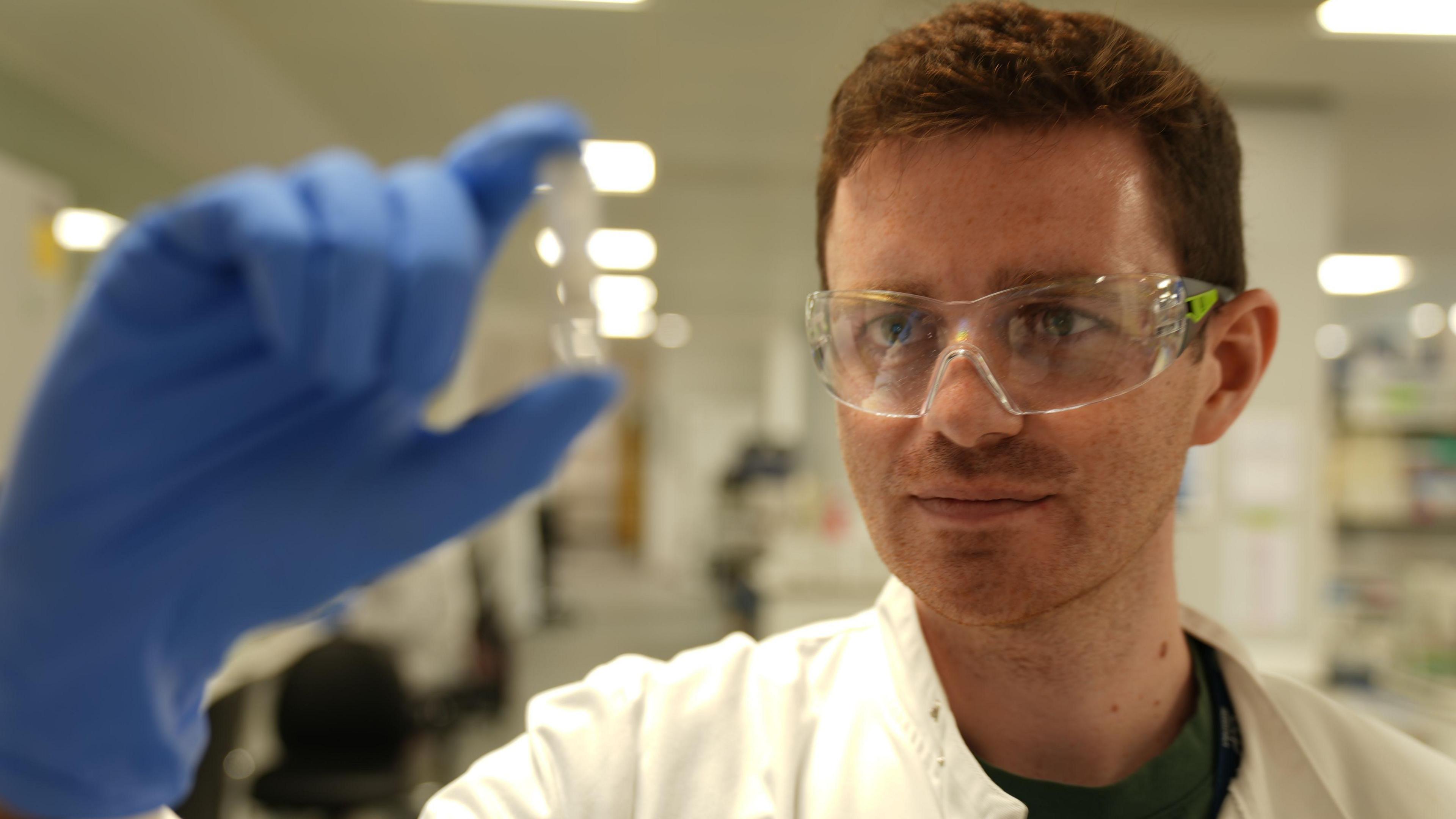 The image shows a man in a laboratory wearing a white lab coat, clear protective safety glasses, and a blue glove. The gloved hand is held up in the foreground, pinching a small transparent container or vial, which is slightly out of focus due to the shallow depth of field. The person is positioned on the right side of the frame and appears to be closely examining the object being held up. The background contains bright overhead lights, laboratory furnishings, and equipment, all blurred to emphasize the subject and the item being inspected.
