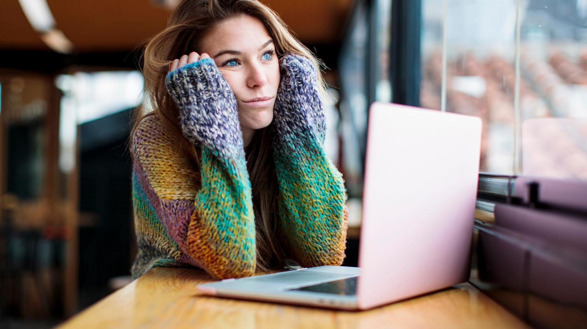 A bored young woman stares out of the window. She is sat in a library, using a laptop