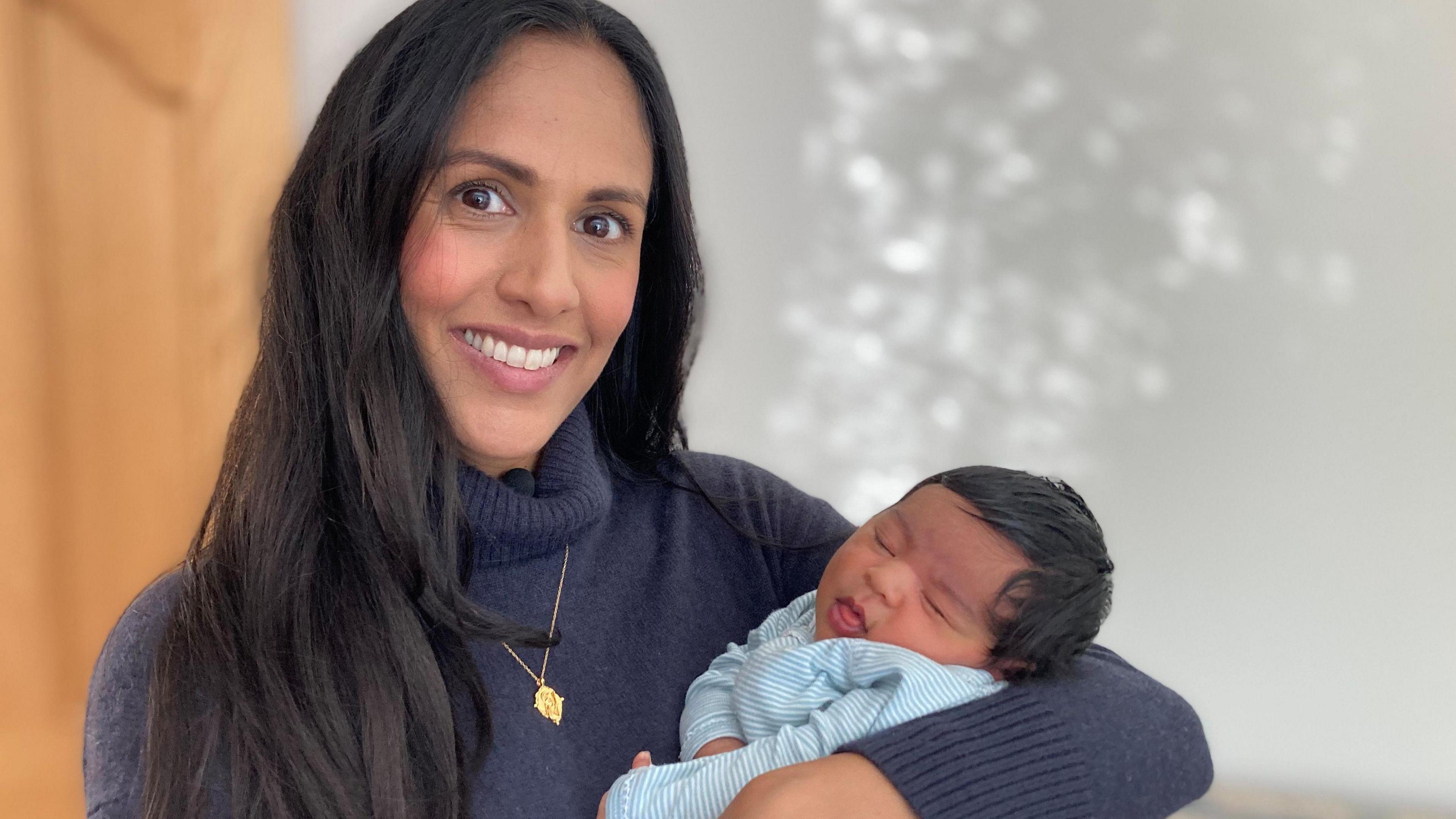 Jasmeen Basi wears a navy blue jumper and is holding her newborn baby in her arms. Jasmeen has long, dark hair and is smiling at the camera. Her baby appears to be asleep.