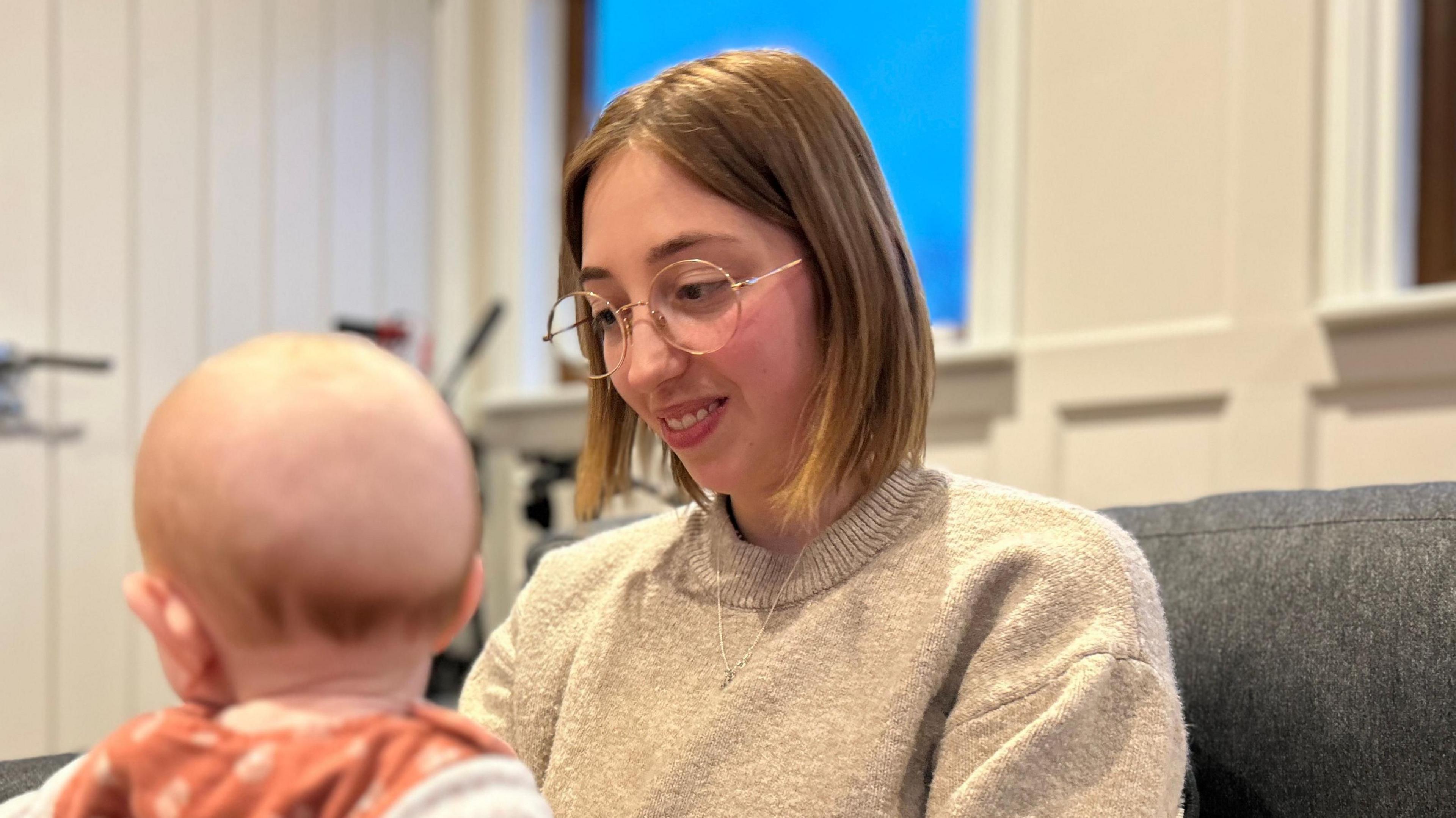 a woman with spectacles and brown hair in a bob sits and looks at her baby. The baby's face is not visible as the image is taken from the back.