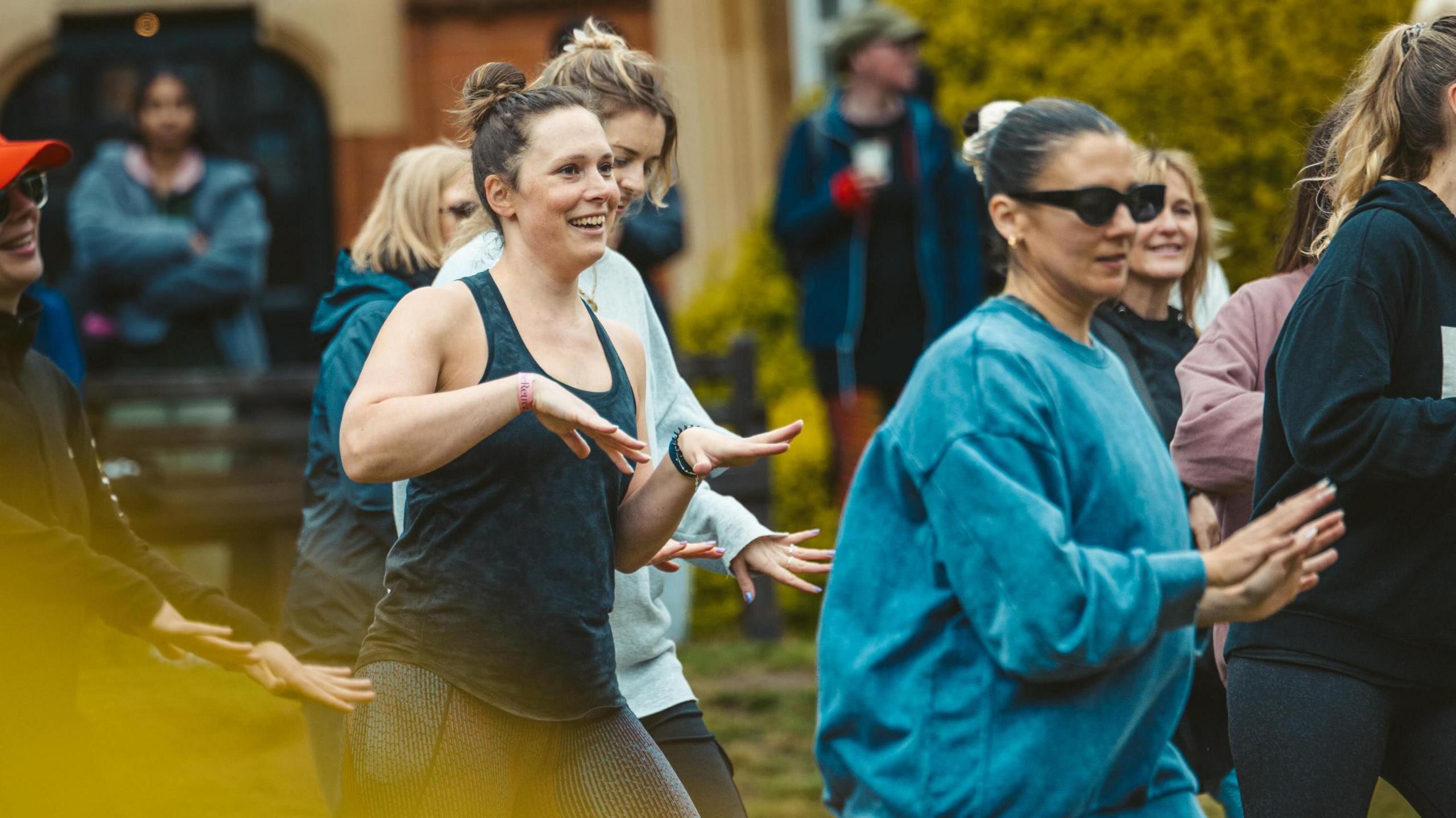 A woman with her hair tied back in a bun is wearing a blue tank top and moving her hands in the air. She is next to other women who are also dancing outside.